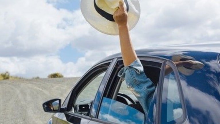 A happy traveler in a moving car raises their hand out the open window, holding a hat in the wind.